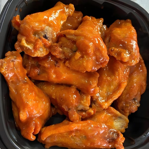 A black container filled with several bright orange, saucy chicken wings from Selfie Subs Food Truck.