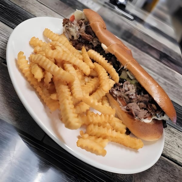 A Selfie Subs cheesesteak sandwich with crinkle-cut fries on a white plate.